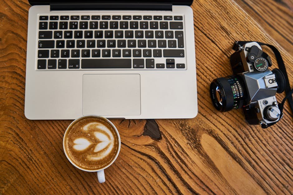 Flat lay of a laptop, cappuccino, and camera on a wooden surface, ideal for work-from-home themes.