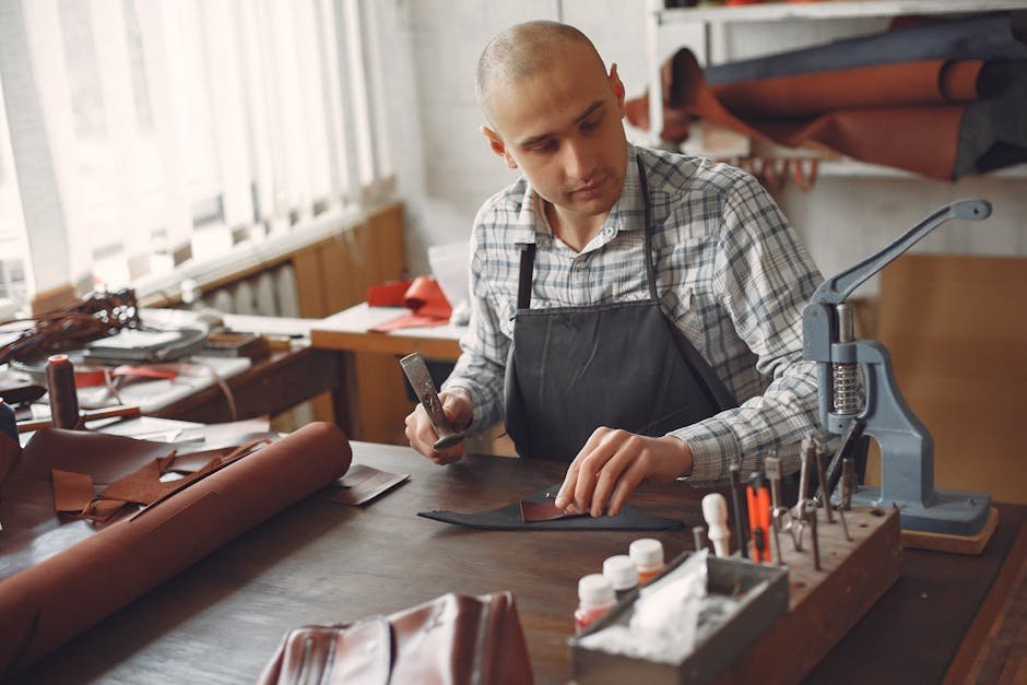 Concentrated adult male artisan in casual shirt and apron using leather piece and hammer while working in workshop