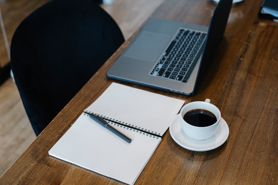 A top view of a modern workspace featuring a laptop, notebook, pen, and coffee cup on a wooden table.