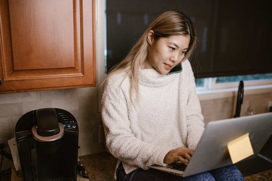 Woman multitasking on laptop and phone in a warm kitchen setup.