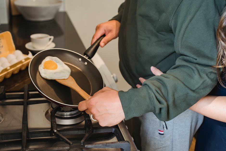 Crop anonymous female embarrassing man in casual clothes standing near gas stove with pan in hand and cooking fried egg for diner in kitchen at home